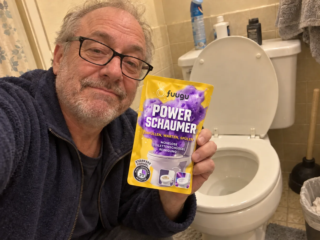 Handy-Selfie eines grauhaarigen Mannes mit Brille, der in einem beige gefliesten Bad einen Fuugu Power Foamer-Beutel neben seine Toilette hält